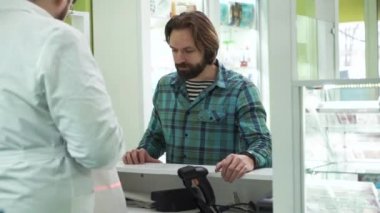 Tişört giymiş sakallı yetişkin beyaz adam eczacının ilaçlarını hazırlamasını bekliyor. Eczacı müşterisine hizmet ediyor. Eczanede alım süreci.