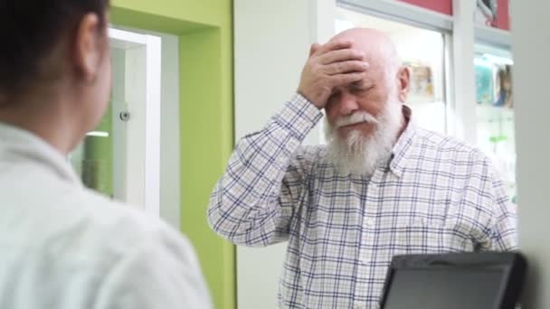 Elderly bearded man with headache coming to the pharmacy. Young female pharmacist offering the customer pills for the common cold. Buyer looking at the unknown medication and refusing to buy it. — Stock Video