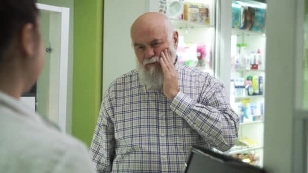 Uomo anziano barbuto con mal di denti che viene in farmacia. Giovane farmacista donna che offre al cliente antidolorifici. Acquirente esaminando il farmaco sconosciuto prima dell'acquisto . — Video Stock