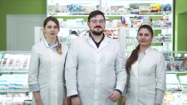 Tres jóvenes farmacéuticos caucásicos mostrando pulgares hacia la cámara y sonriendo. Empleados altamente profesionales que permanecen en su lugar de trabajo. Personas con túnicas blancas destinadas a rescatar vidas . — Vídeo de stock