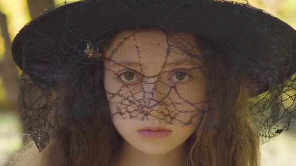 站在秋天公园里的一个戴着万圣节女巫帽的红头发高加索女孩的特写镜头 卷发和棕色眼睛的孩子严肃地看着摄像机 — 图库视频影像