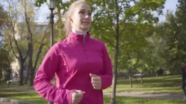 Pembe spor kıyafetleri ve sonbahar parkında çalışan kulaklıklı, gülümseyen beyaz bir kızın yakın çekimi. Sabah açık havada bayan koşucu eğitimi. Spor konsepti