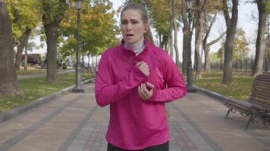 Göğüs ağrısı yüzünden koşuda duran pembe spor giysili çekici beyaz kadın. Bayan koşucu sonbahar parkında sabah antrenmanında kendini kötü hissediyor. Spor ve sağlık konsepti.