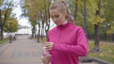Pembe spor giyimli beyaz bir kadının portresi kameraya yaklaşıyor, su içiyor ve öne doğru koşuyor. Sonbahar parkında sabahları kendine güvenen kadın koşucu eğitimi. Spor konsepti.