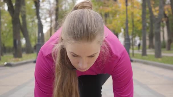 Close-up da jovem mulher caucasiana concentrada na posição inicial olhando para a câmera e começando a correr. Treinamento atraente feminino corredor na parte da manhã no parque de outono. Câmera zooms em . — Vídeo de Stock