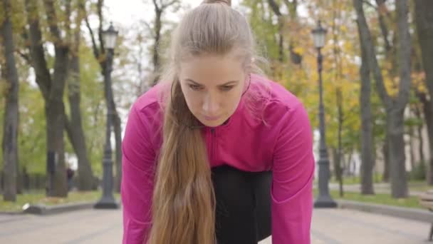 Gros plan de la jeune caucasienne confiante en position de départ regardant vers le haut et commençant à courir. Entraînement attrayant des coureuses le matin dans le parc d'automne. La caméra zoome . — Video