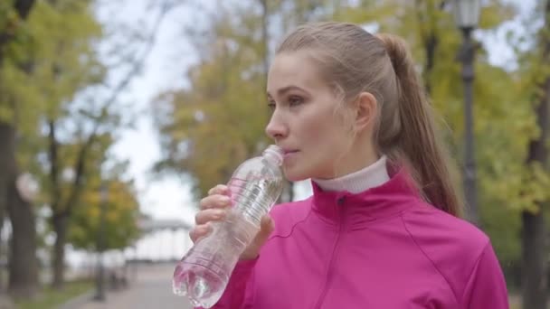 Close-up portrait of a young Caucasian woman in pink sportswear drinking water and running away. Confident female runner training in the autumn park in the morning. Sports concept. — ストック動画