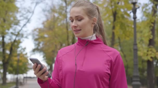 Młoda biała kobieta w różowej odzieży sportowej włącza muzykę w słuchawkach i zaczyna biegać. Żeński trening biegaczy rano na świeżym powietrzu. Koncepcja sportu. — Wideo stockowe