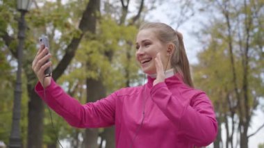 Pembe spor giysili, gülümseyen beyaz kadın portresi video kaset aracılığıyla konuşuyor ve başparmağını gösteriyor. Sabahları açık havada neşeli kız eğitimi. Spor konsepti.