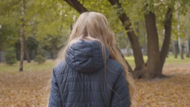 Menina adolescente caucasiana andando no parque de outono, voltando-se e olhando para a câmera. Adolescente bonito em óculos passar tempo livre ao ar livre . — Vídeo de Stock
