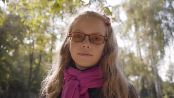 一个年轻的白人女孩站在秋天公园的阳光下 看着相机的肖像 金发碧眼的孩子戴着眼镜在户外休息 — 图库视频影像
