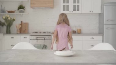 Pembe elbiseli beyaz bir kız arka planda tabak altlığı alıp masaya koyuyor. Çalışkan genç modern mutfakta ev işlerine yardım ediyor.