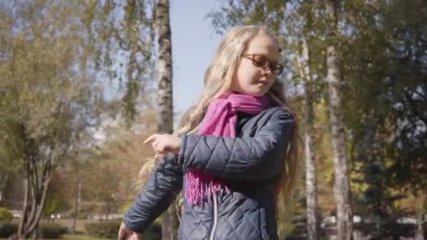 秋の公園や森の木々の間に長いブロンドの巻き毛を持つ陽気な白人の十代の少女 週末は眼鏡をかけて楽しむ女子高生 — ストック動画