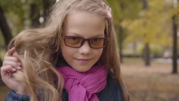 笑逐颜开的白人少女摸着她长长的卷曲的金发 看着相机 在秋天的公园或森林里 身穿光色眼镜和粉色围巾的年轻女学生摆姿势 — 图库视频影像