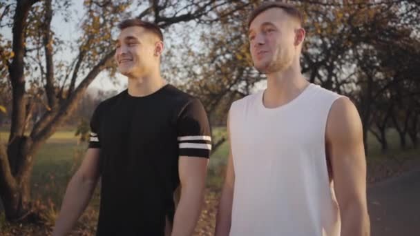 两个穿着运动服的英俊的白人男孩站在秋天公园的阳光下聊天 健美的男人在室外训练后休息 健康生活方式体育概念 — 图库视频影像