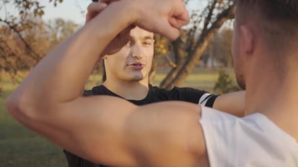 在秋天的公园里 一个年轻的白人运动员在阳光下炫耀自己的肌肉 年轻的健美运动员和他的教练在户外度过了阳光灿烂的一天 健康生活方式概念 — 图库视频影像
