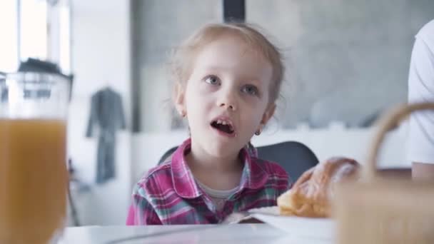 Een close-up portret van een blank meisje dat chocolade eet aan tafel. Gelukkig blond kind met vlechtjes die 's morgens thuis zoet dessert kauwen. Vreugde, vrije tijd, geluk, levensstijl. — Stockvideo