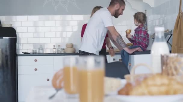 Vrolijke blanke ouders die praten met een schattig meisje op het aanrecht in de keuken. Een breed shot van gelukkige jonge familie die 's ochtends samen thuis zijn. Vreugde, vrije tijd, weekenden. — Stockvideo