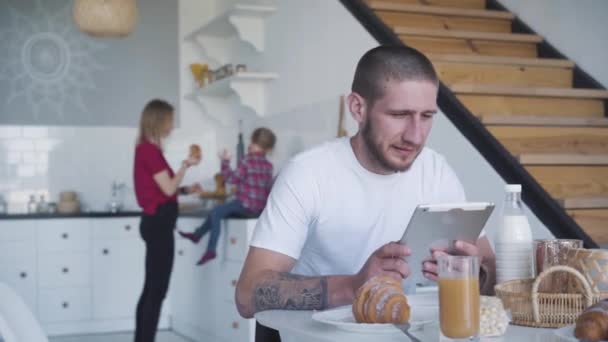 Portrét kavkazského mladého podnikatele, který používá tablet jako blond ženu a malou holčičku připravující snídani v pozadí. Mladý zaneprázdněný muž čeká na rodinu u stolu. Životní styl. — Stock video