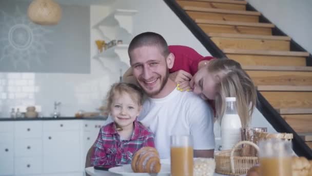 Joyeux jeune famille posant dans la cuisine à la maison. Portrait d'un jeune homme brune souriant, d'une femme blonde et d'une jolie petite fille assise à la table et regardant la caméra. Mode de vie, joie, bonheur, unité . — Video