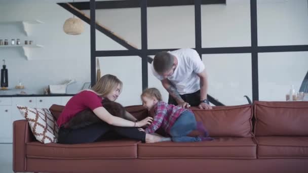 Porträtt av den leende kaukasiske mannen som beundrar sin fru och dotter som leker på soffan. Glad blond kvinna och liten söt flicka pratar som sitter på soffan som far står bakom. Familjetid. — Stockvideo