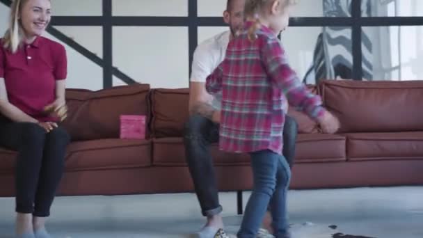 En liten flicka som går i högklackat och klättrar på soffan. Skrattande ung mor och far beundrar sin dotter ha kul hemma. Fritid, glädje, livsstil, lycka, barndom. — Stockvideo