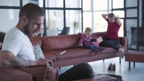 集中注意力的年轻白人男子在周末用平板电脑作为模糊的家庭休息的背景。重点纹身的家伙无视妻子和女儿在沙发上玩耍。生活方式，注意. — 图库视频影像
