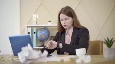 Tükenmiş beyaz kadın boş kahve fincanını kontrol ediyor ve klavyede daktilo yazıyor. İnternette çalışan yorgun beyaz kadın serbest çalışanın portresi. İş, uzak iş, yaşam tarzı.