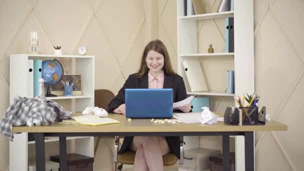 La cámara se acerca a la joven pelirroja sonriente sentada en una habitación desordenada frente a la computadora portátil y hablando. Retrato de freelancer caucásico trabajando desde casa. Estilo de vida, negocios . — Vídeo de stock