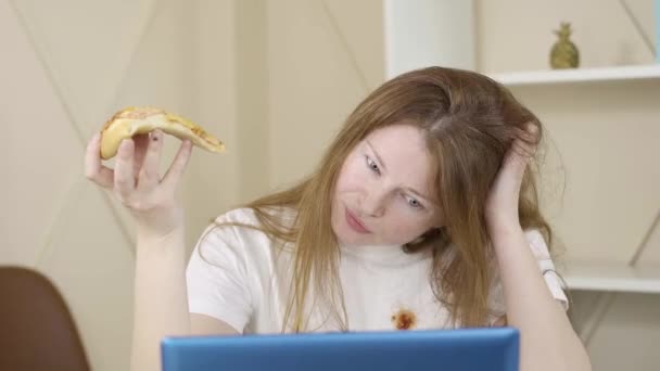 더러운 티셔츠를 입은 여성 프리랜서가 노트북 앞에서 피자를 먹고 있는 모습. 게으름뱅이 백인빨간 머리 여자가 정크푸드를 가지고 집에서 일하는 것을 클로즈업했습니다. 프리랜서, 사업, 생활 방식. — 비디오