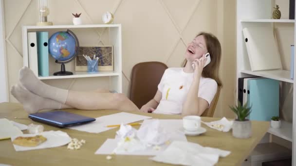 Zrelaksowana, radosna kobieta rozmawiająca przez telefon, jak siedząca przy stole. Portret radosnej białej rudej dziewczyny cieszącej się izolacją w domu w brudnym pokoju. Relaks, lenistwo, styl życia. — Wideo stockowe