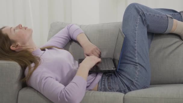 Молодая женщина лежит на диване и печатает на клавиатуре ноутбука. Портрет рыжеволосой белой девушки в интернете или в соцсетях по выходным. Стиль жизни, зависимость от устройств, досуг . — стоковое видео
