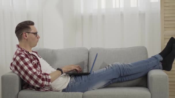 Vista lateral do homem morena deitado no sofá e usando laptop. Ampla foto de cara caucasiano confiante navegando na Internet ou usando mídias sociais. Estilo de vida, freelance, mensagens sem fio e comunicação . — Vídeo de Stock