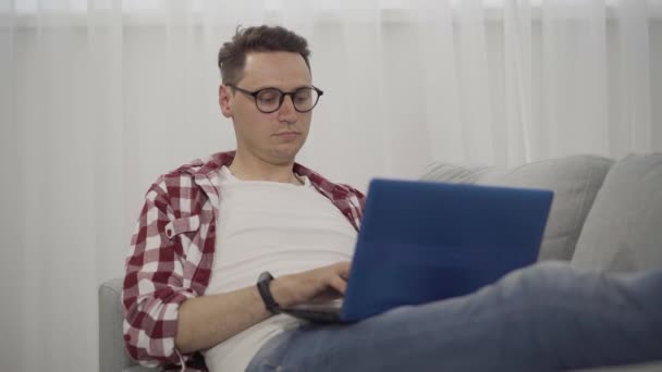 Hombre morena concentrado en anteojos escribiendo en el teclado del portátil. Retrato de una joven morena caucásica navegando por Internet o usando las redes sociales. Estilo de vida, descanso, adicción al dispositivo . — Vídeos de Stock