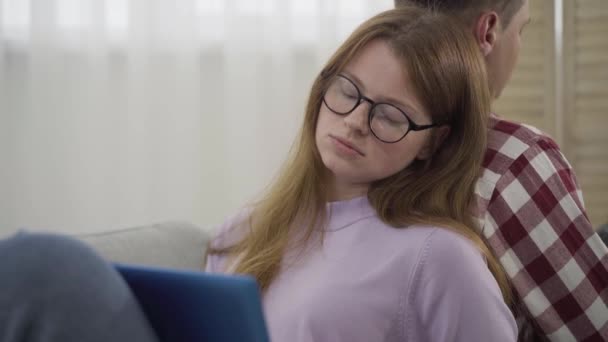 一个疲惫的年轻女人靠在男人的背上打呵欠的画像。戴眼镜的红头发美女白种人在网上用笔记本电脑工作。自由、生活方式、超负荷工作. — 图库视频影像