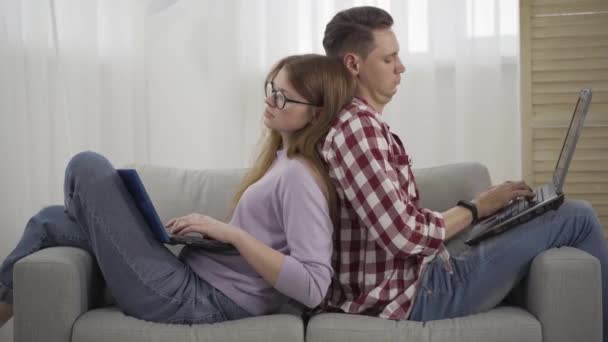Uvolněný tisíciletý pár, který se o sebe opírá na gauči a píše na klávesnici notebooku. Boční pohled portrét absorbovaného mladého bělocha muže a ženy, kteří doma používají sociální média. Životní styl, závislost. — Stock video