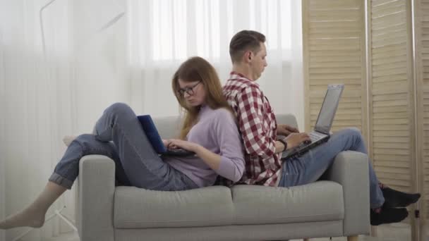 Vista lateral amplia toma de hombre y mujer jóvenes sentados en el sofá se inclinaron el uno al otro y el uso de computadoras portátiles. Millennial Caucásico novio y novia navegar por Internet. Adicción a las redes sociales, estilo de vida . — Vídeo de stock