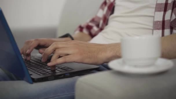 Primer plano de las manos masculinas escribiendo en el teclado del ordenador portátil. Hombre caucásico joven irreconocible tomando taza de café como trabajo en línea en casa. trabajo remoto, freelance, confianza, éxito . — Vídeo de stock