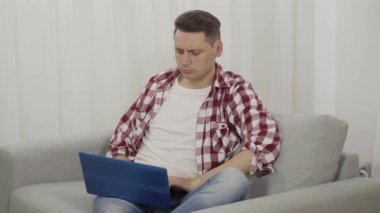 Dizüstü bilgisayarda çalışan yorgun milenyum serbest çalışanı. Kanepede oturup internette sörf yapan yorgun esmer beyaz adamın portresi. Uzaktan çalışma, aşırı çalışma, yaşam tarzı.