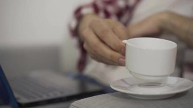 Erkek el kahve fincanı koyuyor ve bulanık klavyeye yazıyor. İnternette çalışan tanınmayan bir adamın yakın çekimi. Serbest konsept, uzak çalışma, yaşam tarzı.