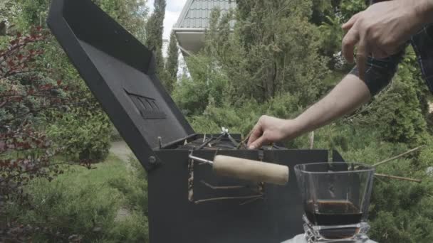 Onherkenbare man steekt bbq op met barbecue rooster. Drinkglas met whisky en cola op de voorgrond. Concept van buiten feesten. kookgelegenheid, levensstijl. — Stockvideo