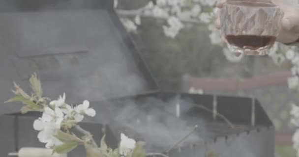 Masculino caucásico mano sosteniendo vidrio con whisky con barbacoa borrosa en el fondo. Hombre adulto irreconocible descansando al aire libre preparando barbacoa. Estilo de vida, relajación, cocina de verano . — Vídeos de Stock