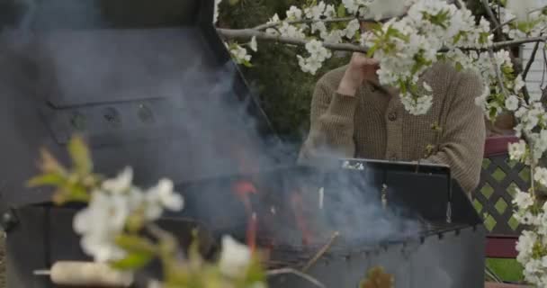 Volwassen blanke man die whisky drinkt achter bloeiende boomtak met wazig brandende bbq grill aan de voorkant. Ontspannen man bereidt barbecue buiten op voorjaars- of zomerdag. Alcohol, feesten, eten.. — Stockvideo