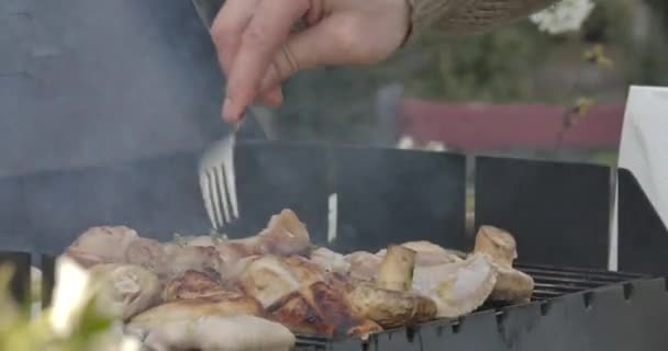 Mężczyzna kaukaski ręka obracając pyszne smażone mięso i grzyby z widelcem. Zbliżenie nierozpoznawalnego mężczyzny smażącego smacznego kurczaka i warzywa na grillu na świeżym powietrzu. Wypoczynek, grillowanie. — Wideo stockowe