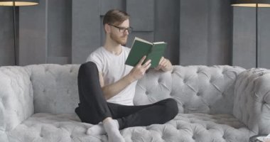 Gözlüklü, evde kitap okuyan, kendinden emin, beyaz bir adam. Genç, yakışıklı, esmer adamın portresi. Kanepede oturuyor ve hobisinin tadını çıkarıyor. İstihbarat. Sinema 4k ProRes Merkezi.