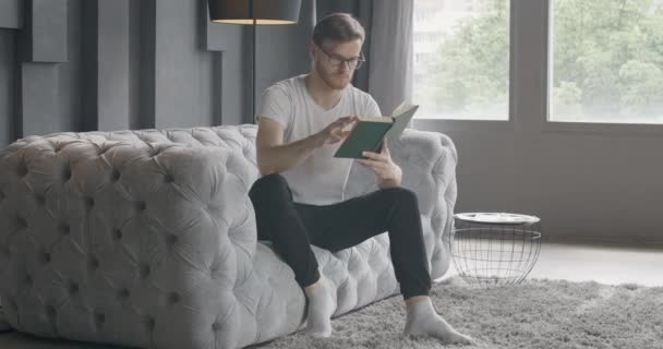 Широкий кадр людини, що перевертає сторінки книги, як сидить на дивані в приміщенні. Портрет поглинаючого молодого кавказького читача, який насолоджується літературою вдома. Читання, хобі, спосіб життя. Фільм 4k ProRes HQ. — стокове відео