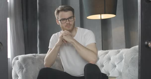 Retrato de hombre morena quitándose las gafas y mirando a la cámara con expresión facial seria. Un hombre caucásico barbudo sentado en el sofá en el interior y pensando. Sede del cine 4k ProRes . — Vídeos de Stock