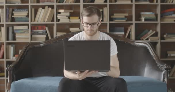 Retrato lento del hombre absorbido serio usando el ordenador portátil en la biblioteca cómoda. Vista frontal de la joven y concentrada morena lectora caucásica sentada al fondo de las estanterías. Sede del cine 4k ProRes . — Vídeo de stock