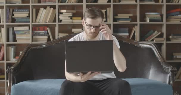 Absorvido jovem sentado com laptop no fundo de estantes e olhando para a tela. Retrato de empresário caucasiano inteligente navegando na Internet na biblioteca. Cinema 4k ProRes HQ . — Vídeo de Stock