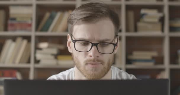 Gözlüklü, dizüstü bilgisayara bakan odaklanmış genç adamın yakın çekimi. Kafkasyalı esmer adamın portresi sosyal medyayı kullanıyor ya da arka planda kitaplıklarla internette sörf yapıyor. Sinema 4k ProRes Merkezi. — Stok video